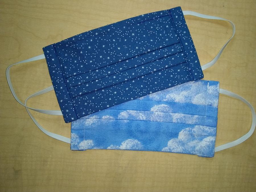 Two pleated face masks with elastic ear loops, lying on wooden background. One in cloud print, one in starts print.