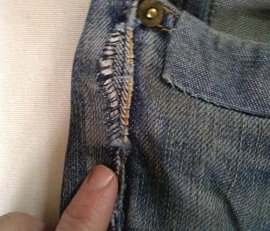 closeup of upper front side of jeans, by pocket, with finger pointing out rip in side seam with rough, preliminary sewing to tack the rip down.