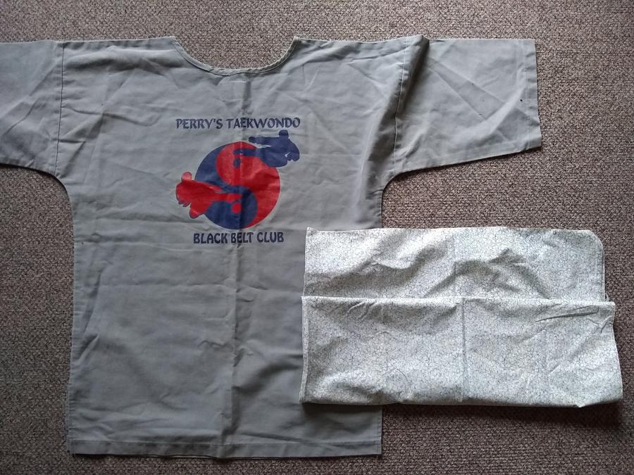 Gray tunic with red and navy martial arts graphic and "Perry's TaeKwonDo Black Belt Club" screen printed on it, with folded up pale printed cotton fabric lying on top of tunic
