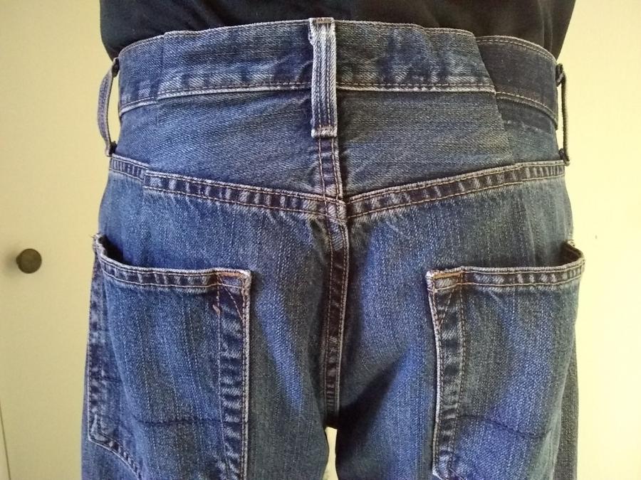 back view of man in blue jeans, from waist to bottom of back pocket, with two darts that slim the waist area visible on either side of beltloop