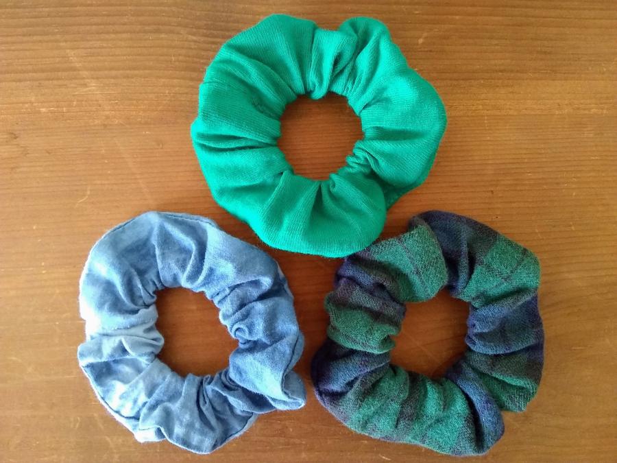 Three scrunchies on a wood surface: blue tie dye, bright green t-shirt material, black watch plaid